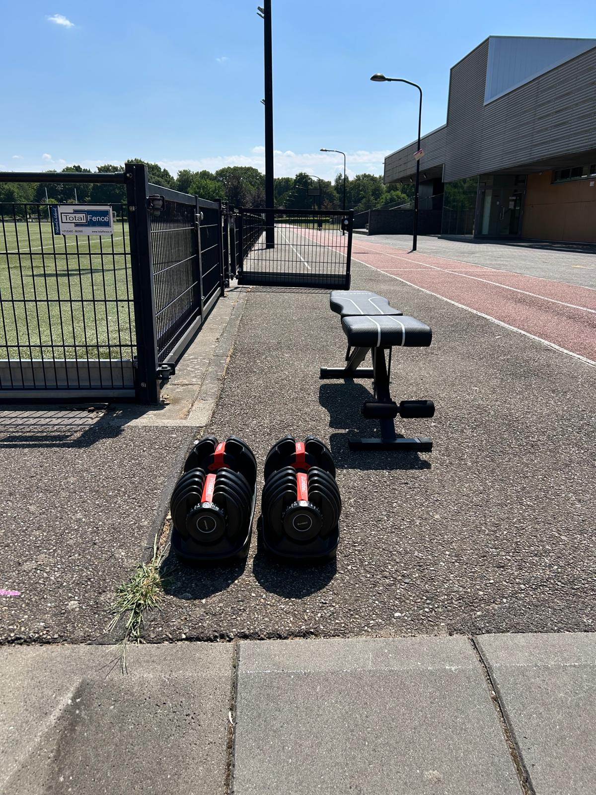 Perform & Relax trainingsmateriaal buiten op de atletiekbaan — dumbbells en bank bij sportveld